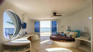 Ein Schlafzimmer mit einer gebogenen Kommode, einem Bett mit vielen Kissen, einem großen runden Spiegel und Blick auf den Strand.