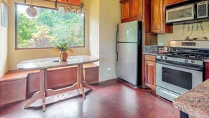 A kitchen with wooden cabinets, a stainless steel refrigerator, a stove, and a window with a view of greenery. - Garden Casita | Pool Access & Mountain Views (Dangriga)