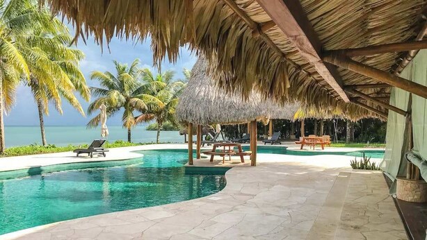 A pool area with a thatched roof, lounge chairs, and a view of the ocean. - Garden Casita | Pool Access & Mountain Views (Dangriga)