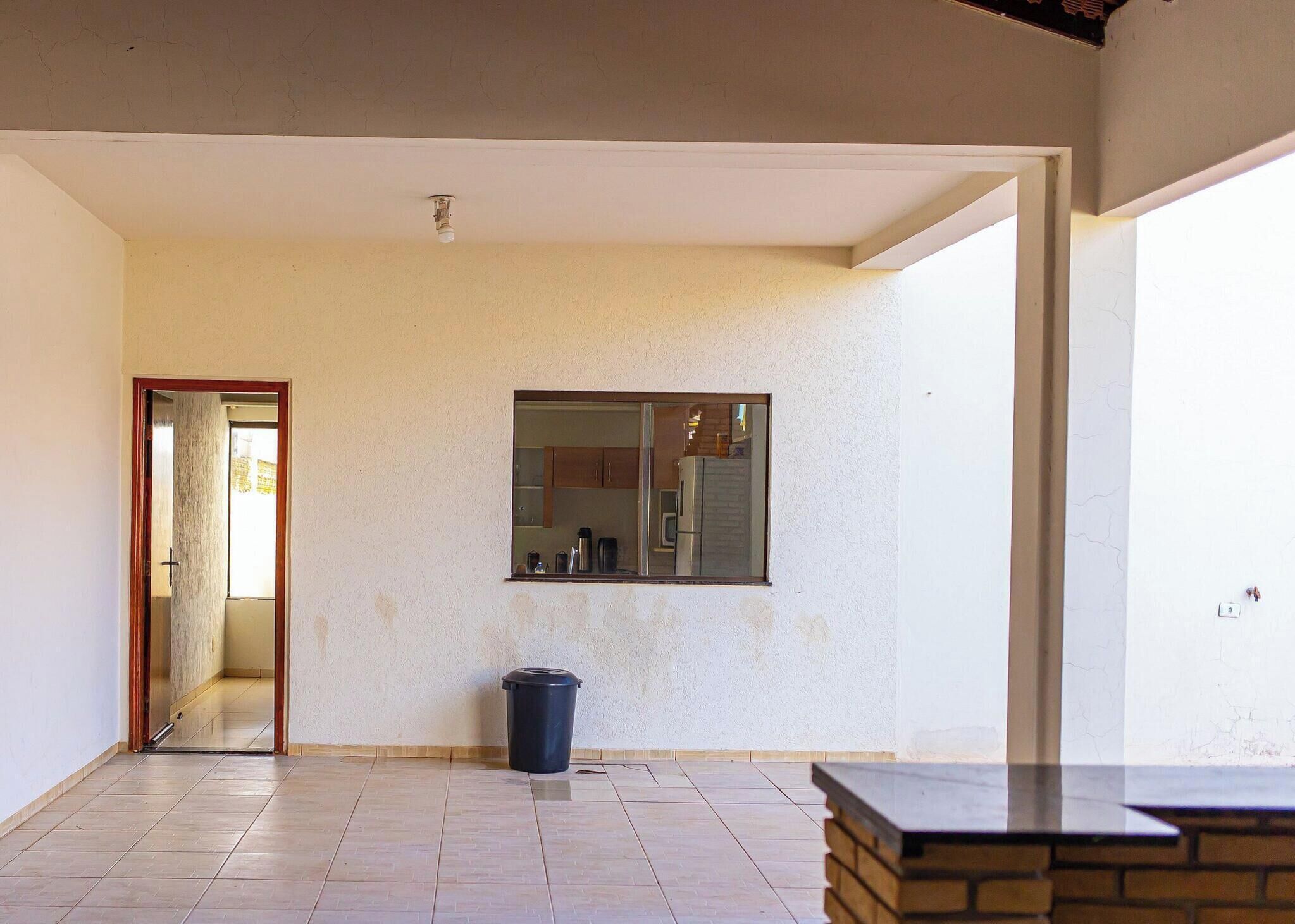 Quarto com piso de azulejos, porta que dá para outro cômodo, lixeira e janela com vista para uma cozinha.