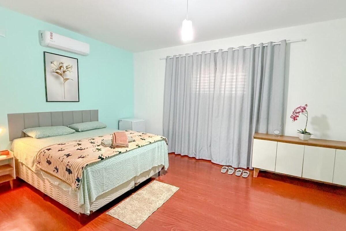 A bedroom with a bed, bedside tables, a nightstand, a dresser, a window with curtains, and a wall-mounted air conditioner.