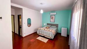 Un dormitorio con una cama, una mesita de noche pequeña, un espejo, un aire acondicionado instalado en la pared y una puerta que da a otra habitación.