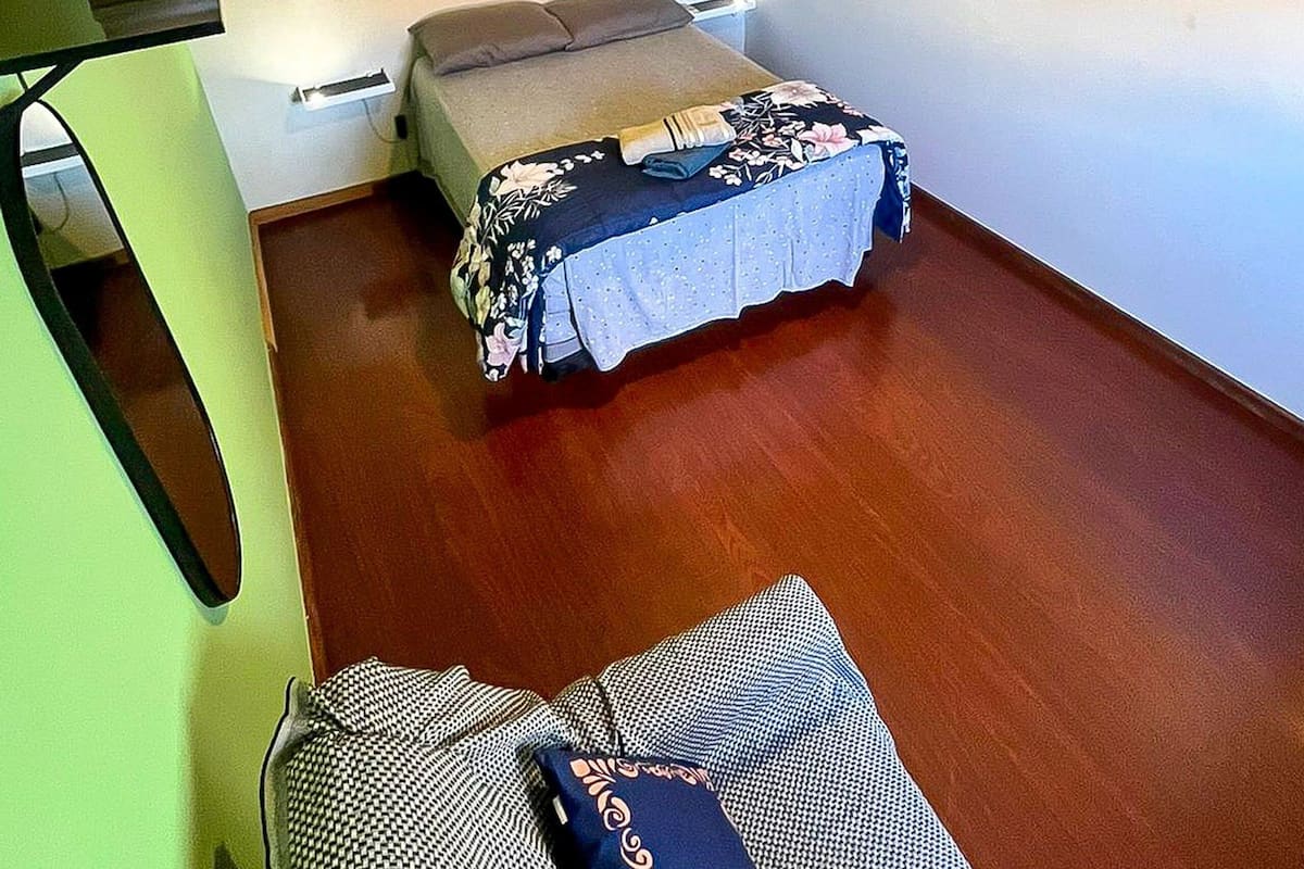 A room with a bed, a bedside table, a wall-mounted shelf with books, and a hanging light fixture.