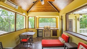 Una stanza con una ventola a soffitto, un tavolo da pranzo con sedie, un angolo cottura con una griglia e ampie finestre che offrono vista sul verde.