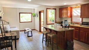 A modern kitchen with wooden cabinets, a central island with stools, a dining area with a sofa and table, and a TV mounted on the wall. - Beachfront abode with well-equipped kitchen - pool, beach, grill, & canoe access (Caye Caulker)