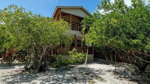 A modern building with a flat roof, surrounded by dense greenery and a sandy ground. - Beachfront abode with well-equipped kitchen - pool, beach, grill, & canoe access (Caye Caulker)