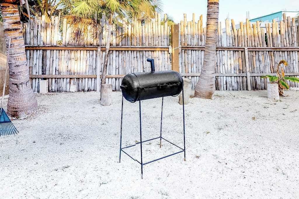 A barbecue grill in a sandy outdoor area with a wooden fence and palm trees.