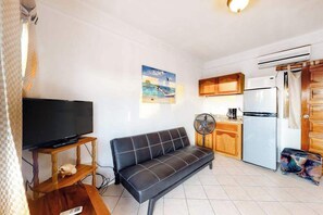 Een woonkamer met een bank, televisie en een keukenhoek, inclusief een koelkast en ventilator.