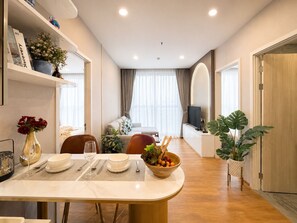 A modern dining area with a white table, chairs, and a bowl of fruit. There is a sofa and a TV in the background. - Centralworldbts (Bangkok)