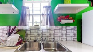 Une cuisine dotée de deux éviers en acier inoxydable, d’une fenêtre ornée de rideaux violets et d’un mur vert agrémenté d’un pare-feu en carreaux à motifs géométriques.