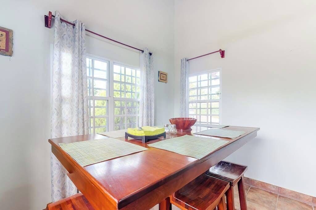 A room with a long wooden table, two windows with curtains, a small stool, and a framed picture on the wall.