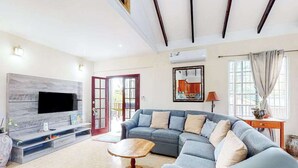 Ein Wohnzimmer mit einem Ecksofa, einem Flachbildfernseher an einer Holzwand, einem Couchtisch aus Holz und einer Tür, die zu einem Außenbereich führt.