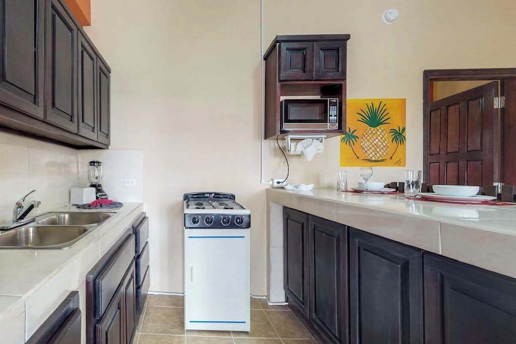 Een keuken met donkere kastjes, een witte kookplaat, een magnetron en een schilderij van ananassen.