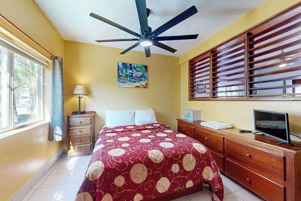 A bedroom with a bed, a ceiling fan, a window with blinds, a desk with a computer, and a TV.
