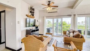 Ein Wohnzimmer mit Rattanmöbeln, einem Deckenventilator, einem Flachbildfernseher und Blick durch große Glasfenster.