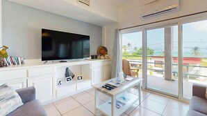 Una sala de estar moderna con un televisor de pantalla plana, un mueble blanco para entretenimiento, una mesa de centro de vidrio y un balcón con vistas.