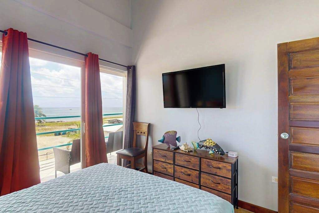 Un dormitorio con una cama, un televisor montado en la pared, un tocador de madera y vista al océano.
