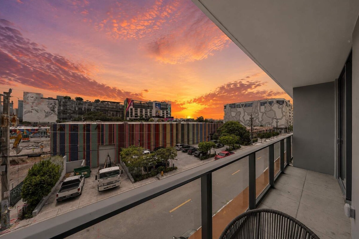Un balcone con vista su un tramonto sopra una città, con edifici e auto parcheggiate.