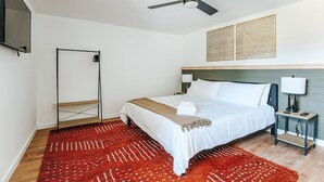 Una habitación de hotel con una cama, una mesita de noche, un televisor, un ventilador de techo y una alfombra roja estampada.