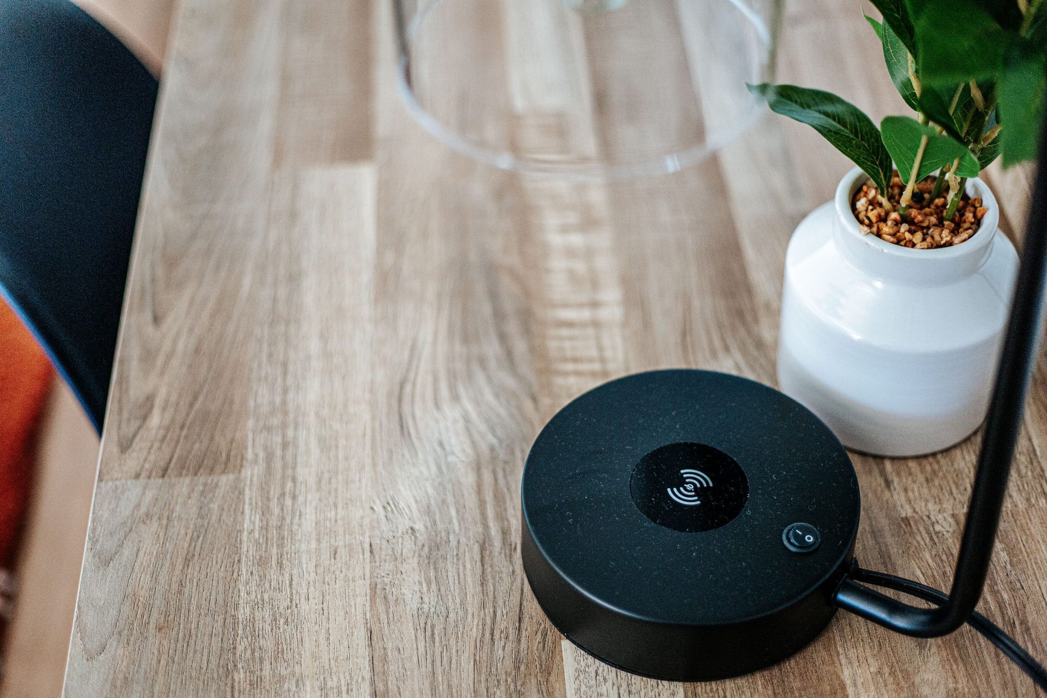 Een rond zwart apparaat met een signaalpictogram op een houten ondergrond, een witte vaas met groene planten, en een glazen object op de achtergrond.