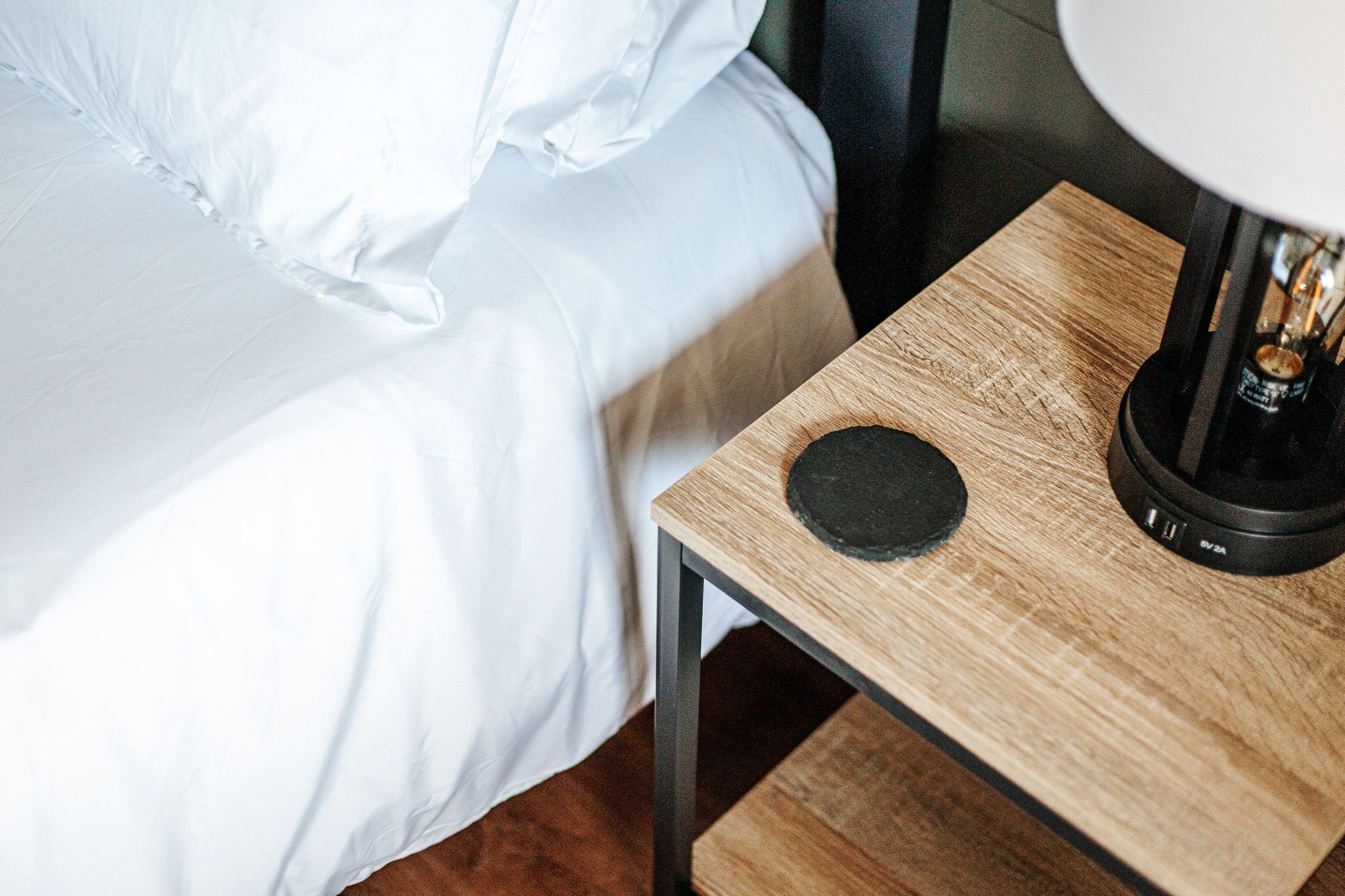 Un lit bien fait, avec du linge de lit blanc, une petite table de chevet en bois sur laquelle se trouve une lampe, et un objet rond posé sur la table de chevet.