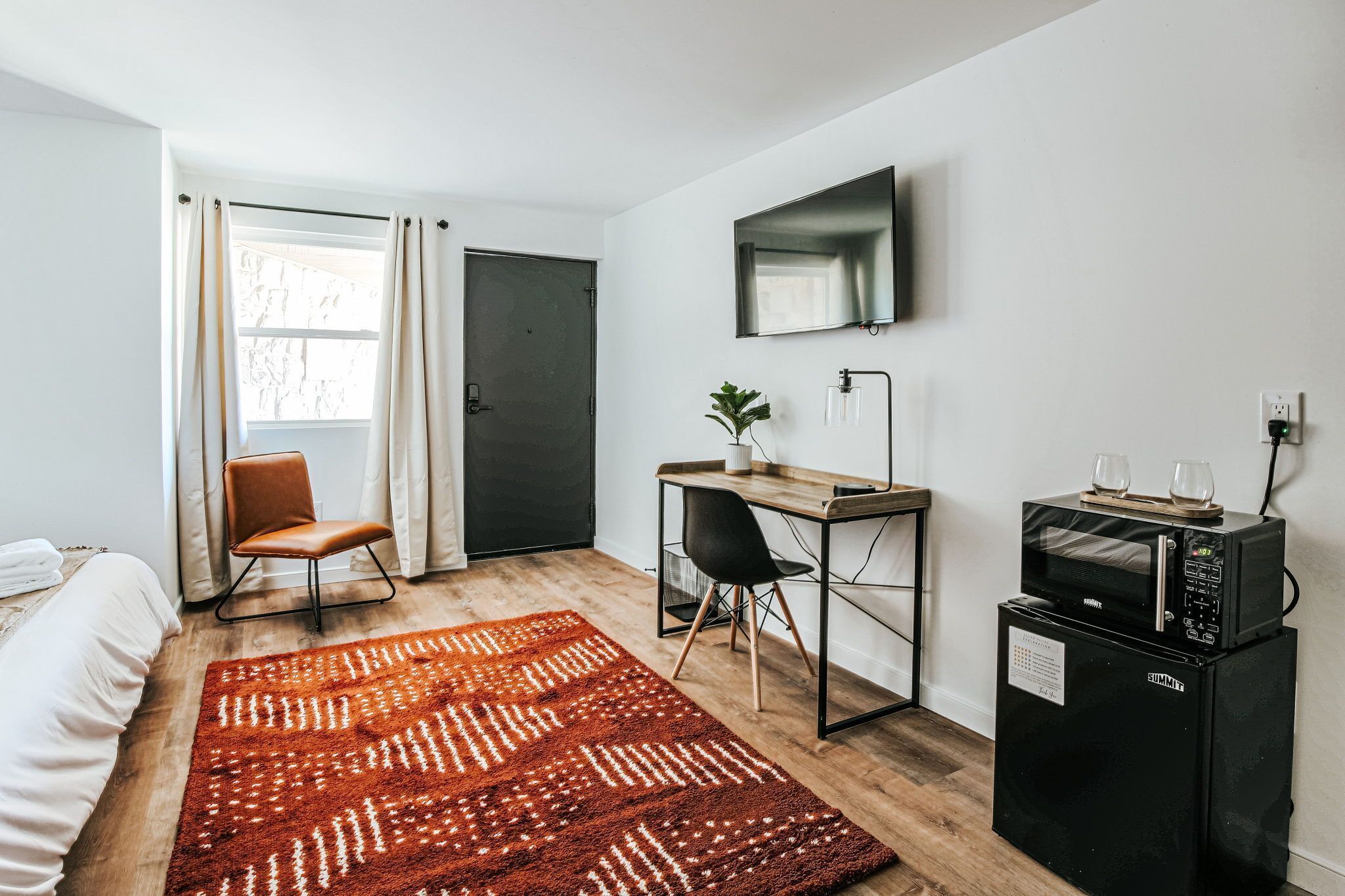 Une chambre d’hôtel compacte comprenant un lit, un bureau avec une chaise, un four à micro-ondes, un petit réfrigérateur, une télévision et une fenêtre avec des rideaux.