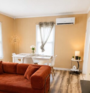 Ein Zimmer mit einer Couch, einem Esstisch mit Stühlen, einem kleinen Tisch mit einer Lampe und einer Klimaanlage an der Wand.