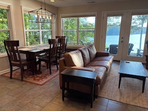 A dining area with a table, chairs, and a sofa. There are large windows with a view of trees and a body of water.