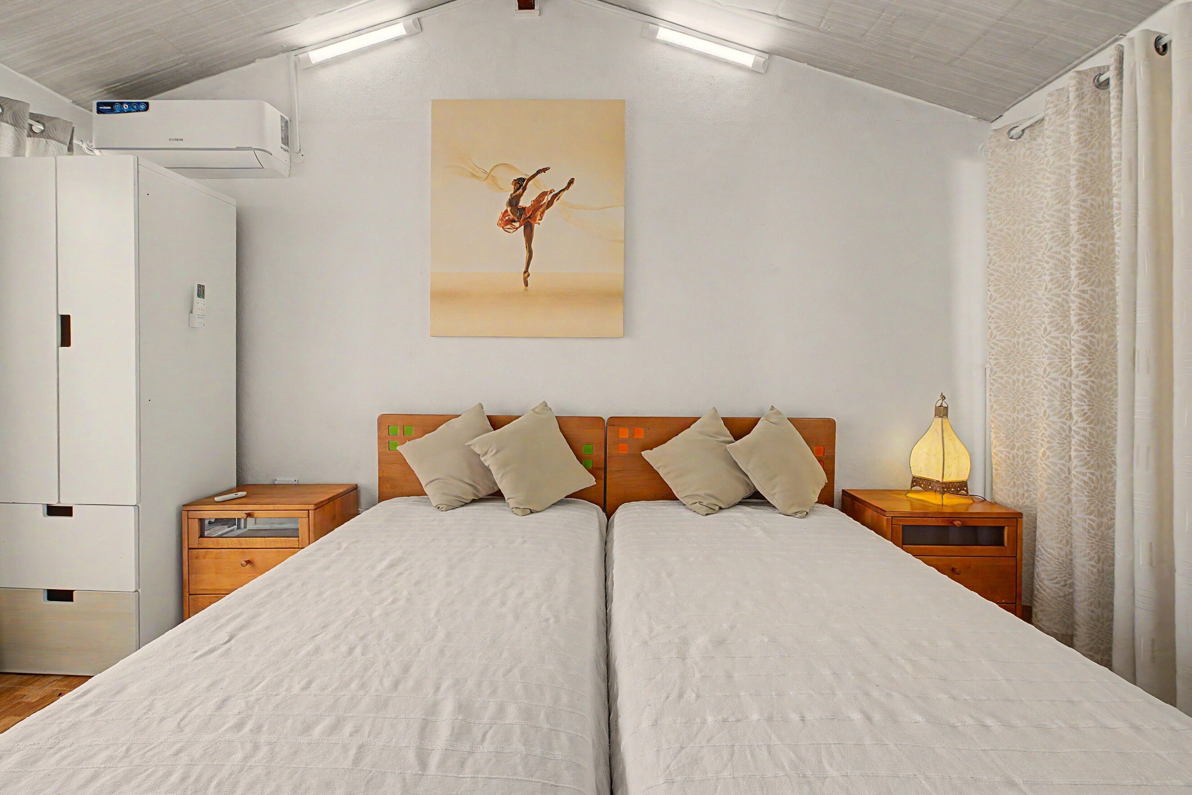 Een tweepersoonsbed met houten hoofdeinde, twee nachtkastjes, een wandairconditioner en een schilderij van een danseres.