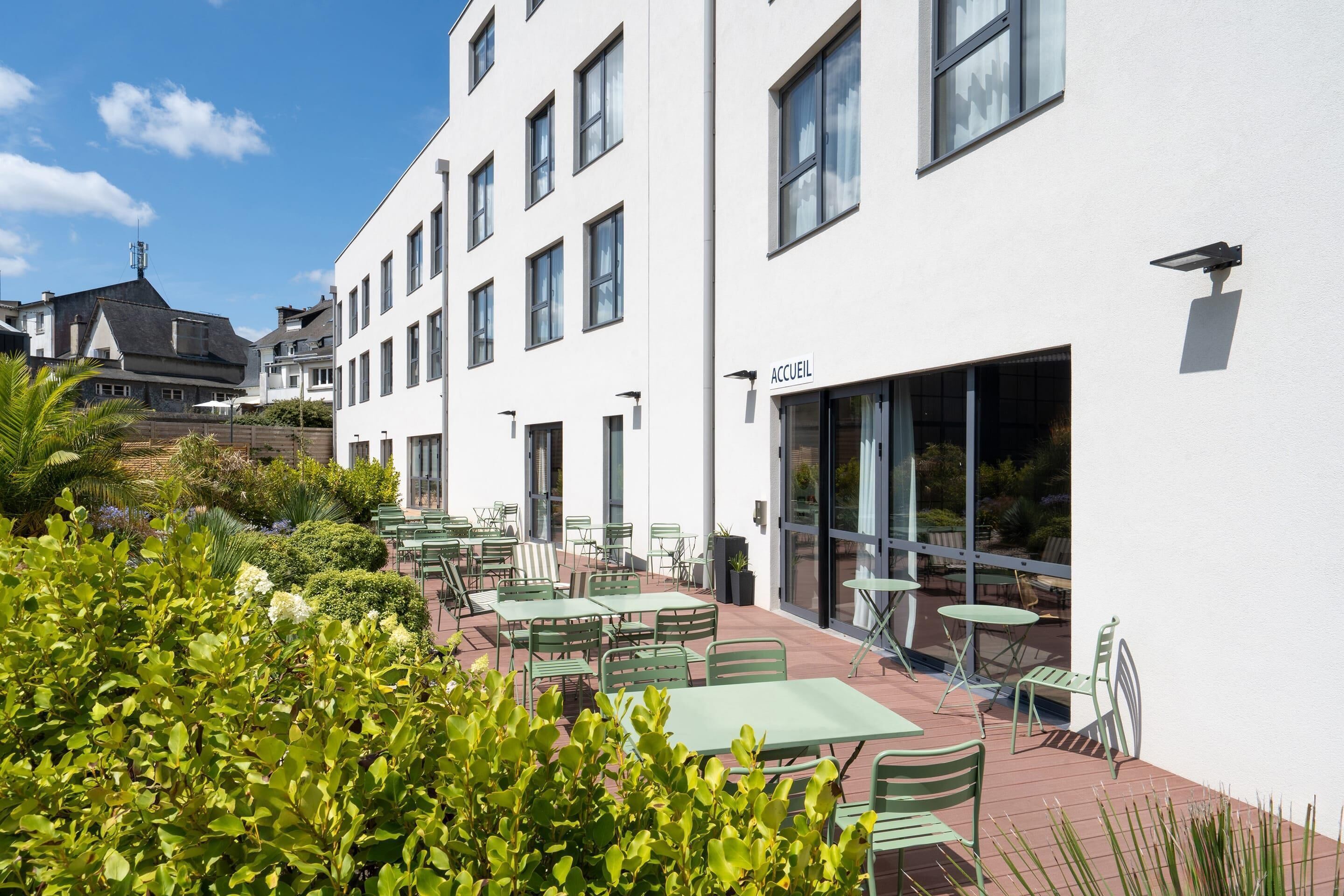 Een hotel met een witte gevel, meerdere ramen en een terras met groene metalen tafels en stoelen.