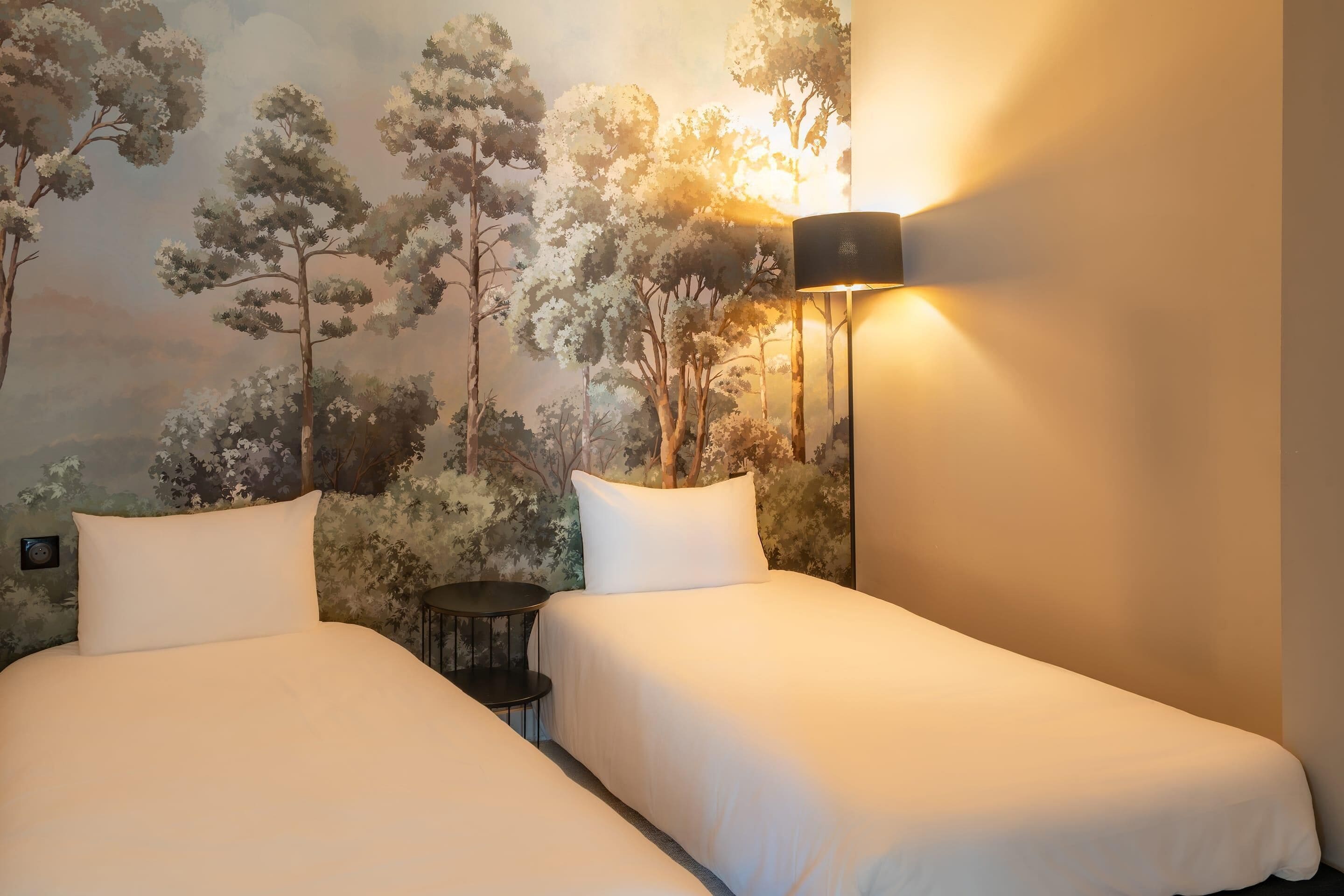 Habitación de hotel con dos camas, una mesa redonda pequeña y un mural en la pared con árboles.