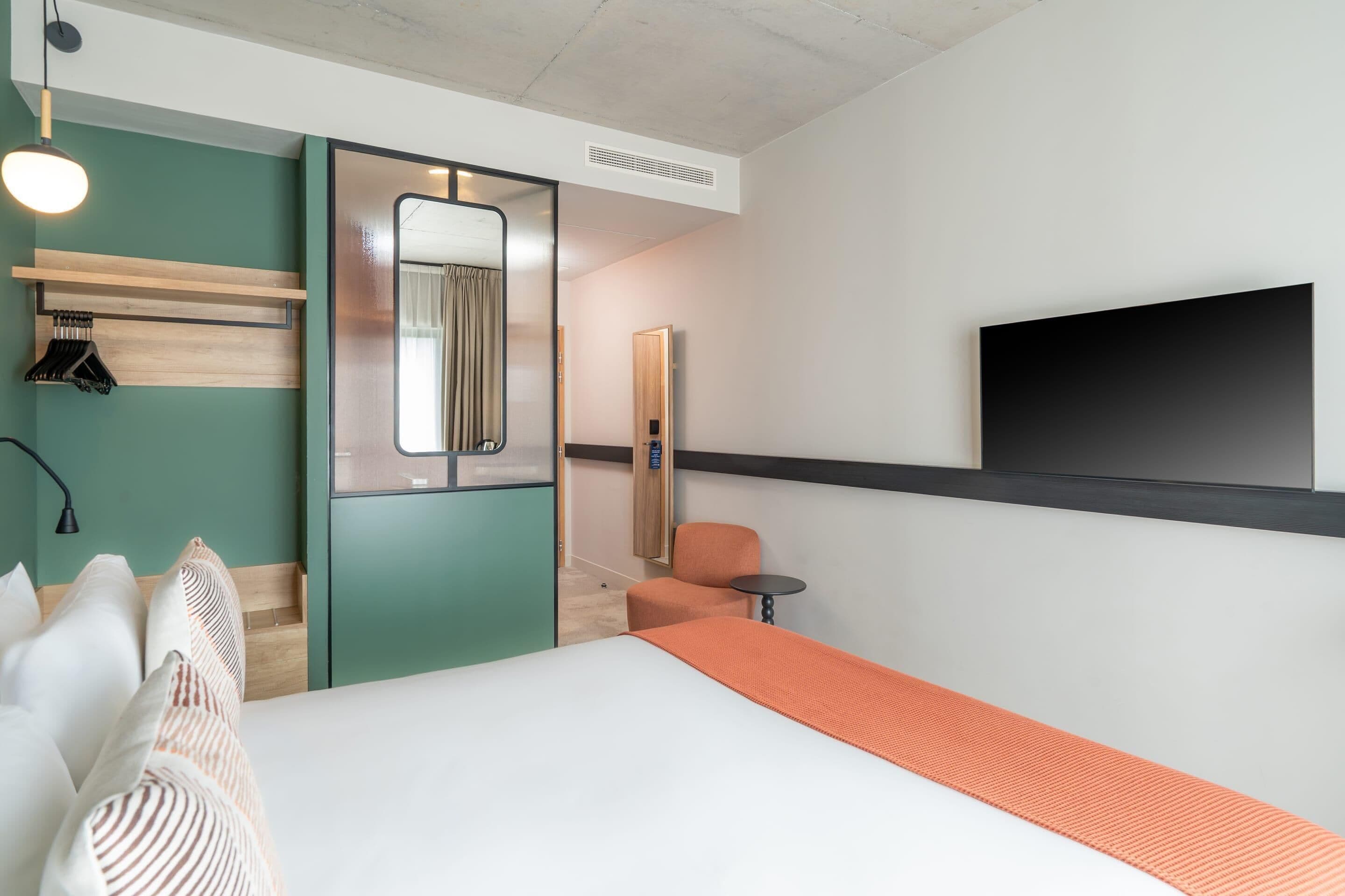 Une chambre d’hôtel comprenant un lit, un coin salon avec un fauteuil et une petite table, un porte-manteau, un miroir et un téléviseur fixé au mur.