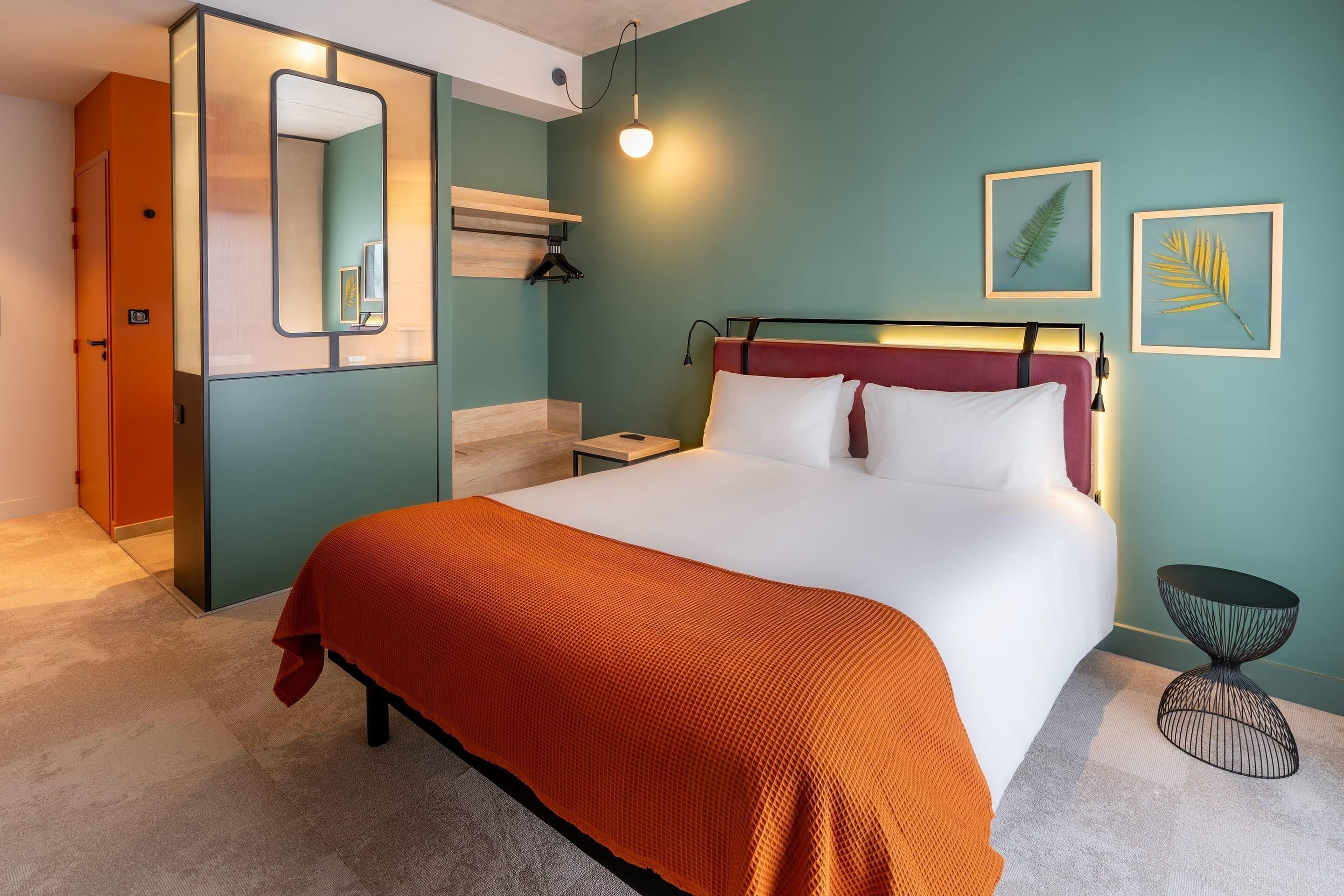 A hotel room with a bed, a bedside table, a nightstand, a mirror, a hanging light, and two framed leaves on the wall.