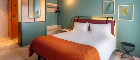 A hotel room with a bed, a bedside table, a nightstand, a mirror, a hanging light, and two framed leaves on the wall.