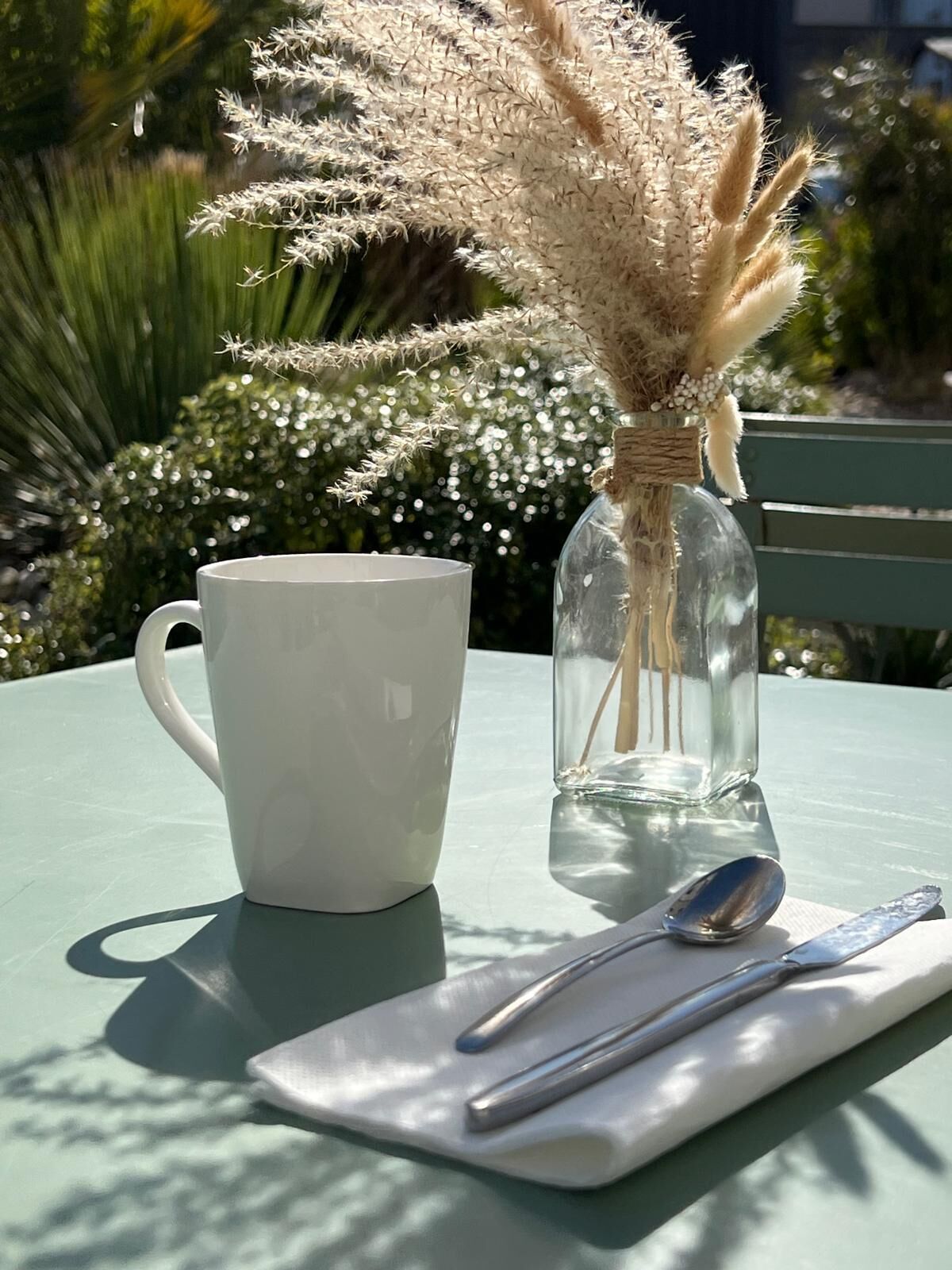 A white mug, a clear glass bottle with pampas grass, and a spoon on a white napkin on a table.