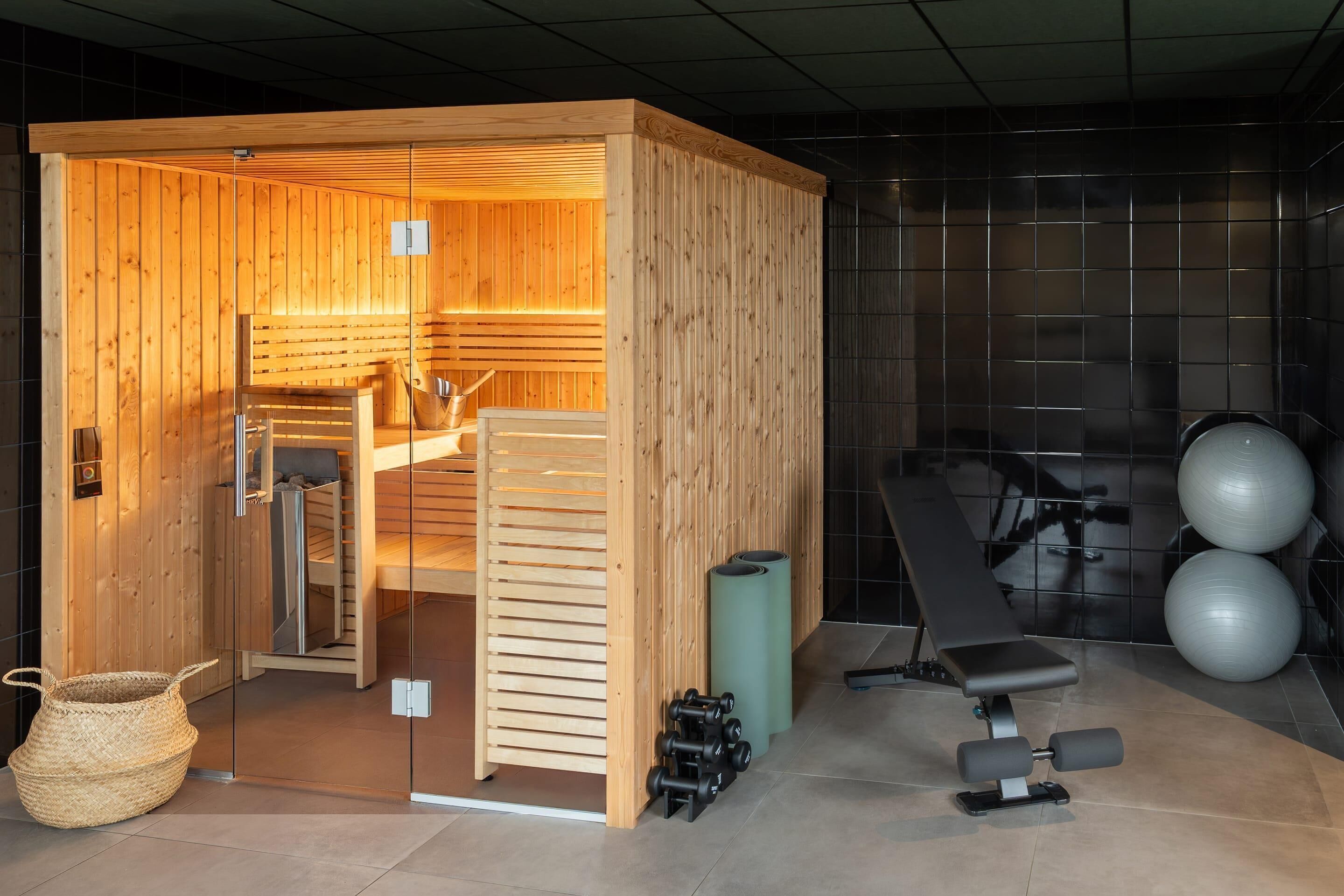Een sauna met houten wanden, een bank, een sauna-oven en fitnessapparatuur, waaronder gewichten en fitnessballen.