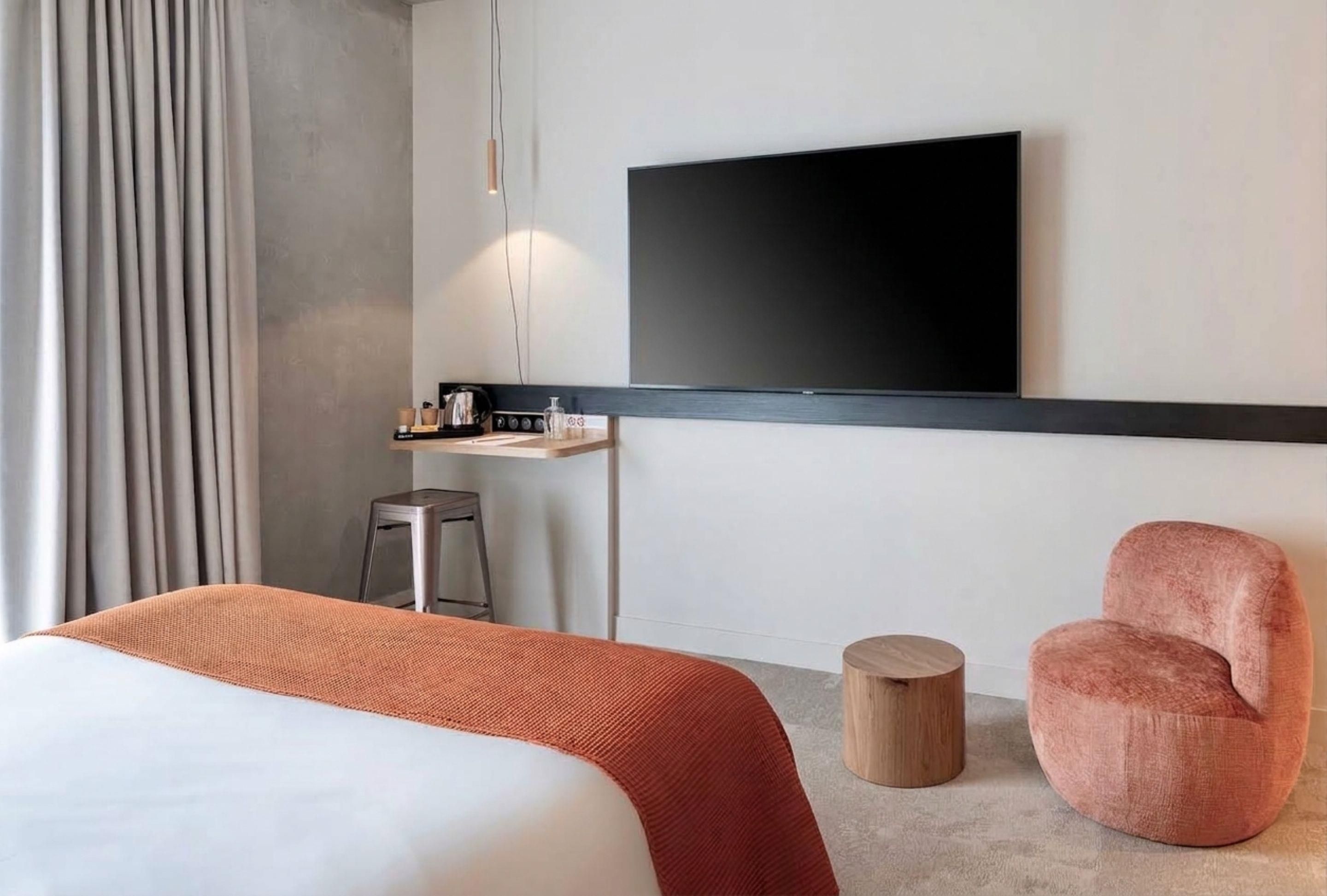 Une chambre d’hôtel moderne avec un grand lit, un téléviseur fixé au mur, un petit bureau avec une chaise et un tabouret rond en bois.