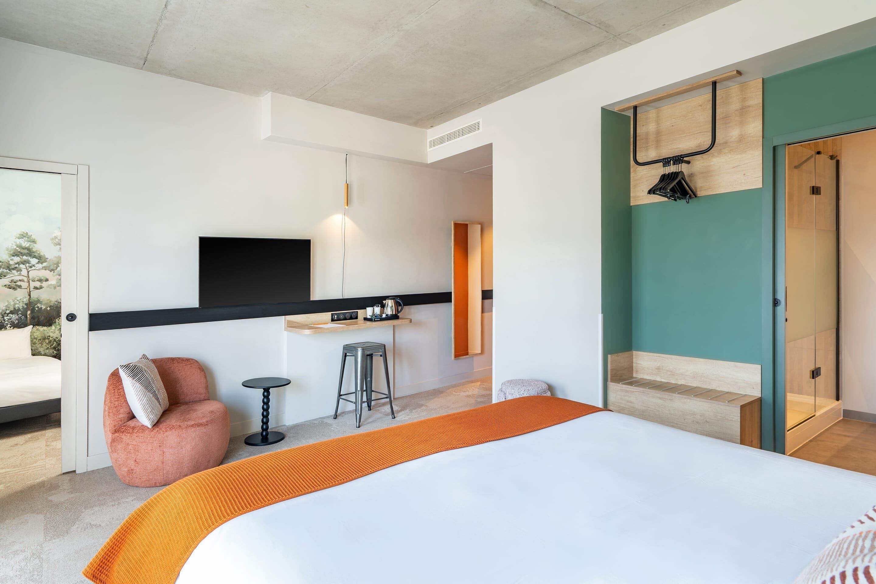 Una habitación de hotel moderna con una cama, un pequeño rincón de cocina, un área de descanso y un televisor montado en la pared.