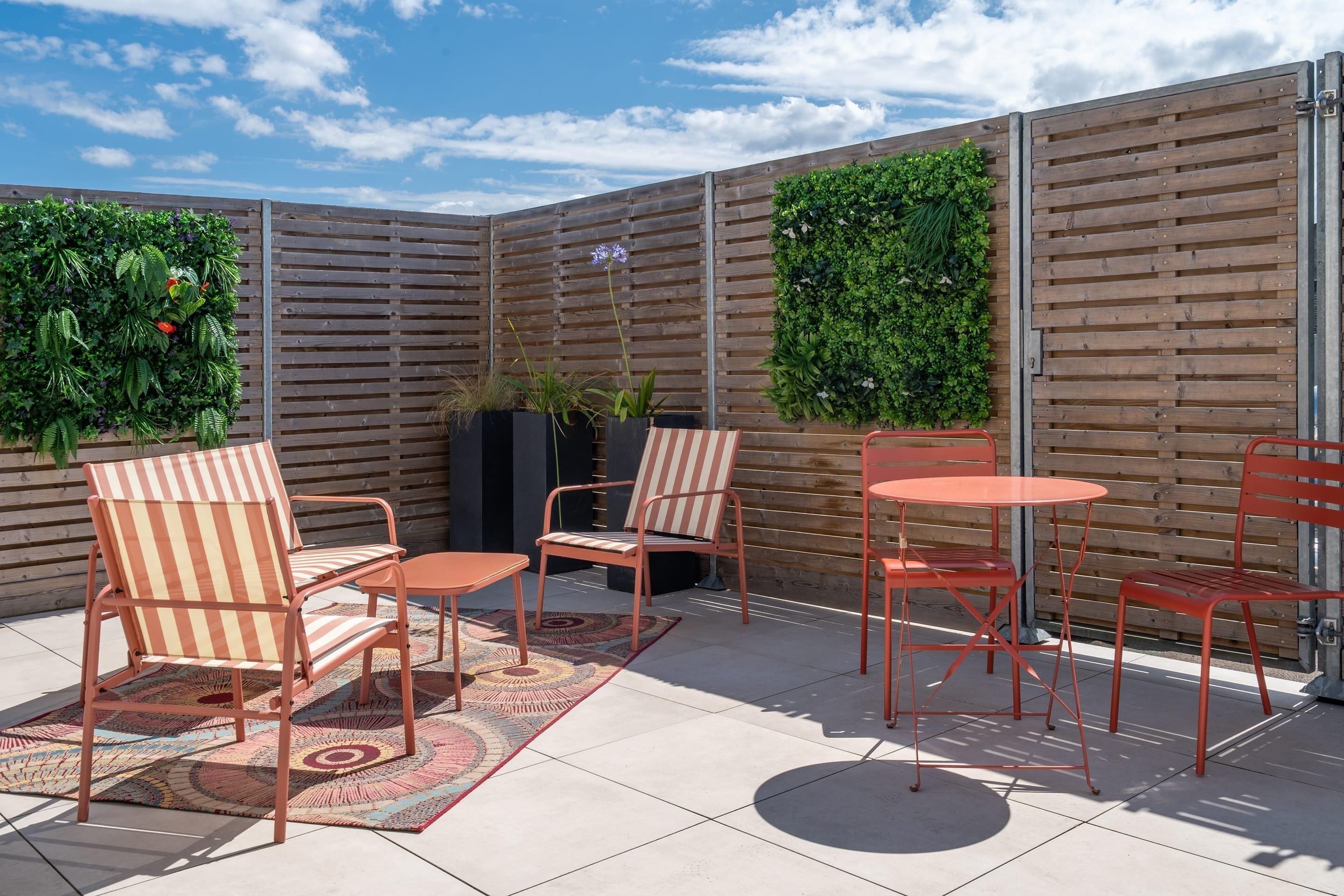 A patio area with a seating arrangement including chairs and a table, surrounded by a wooden fence with vertical slats and greenery.