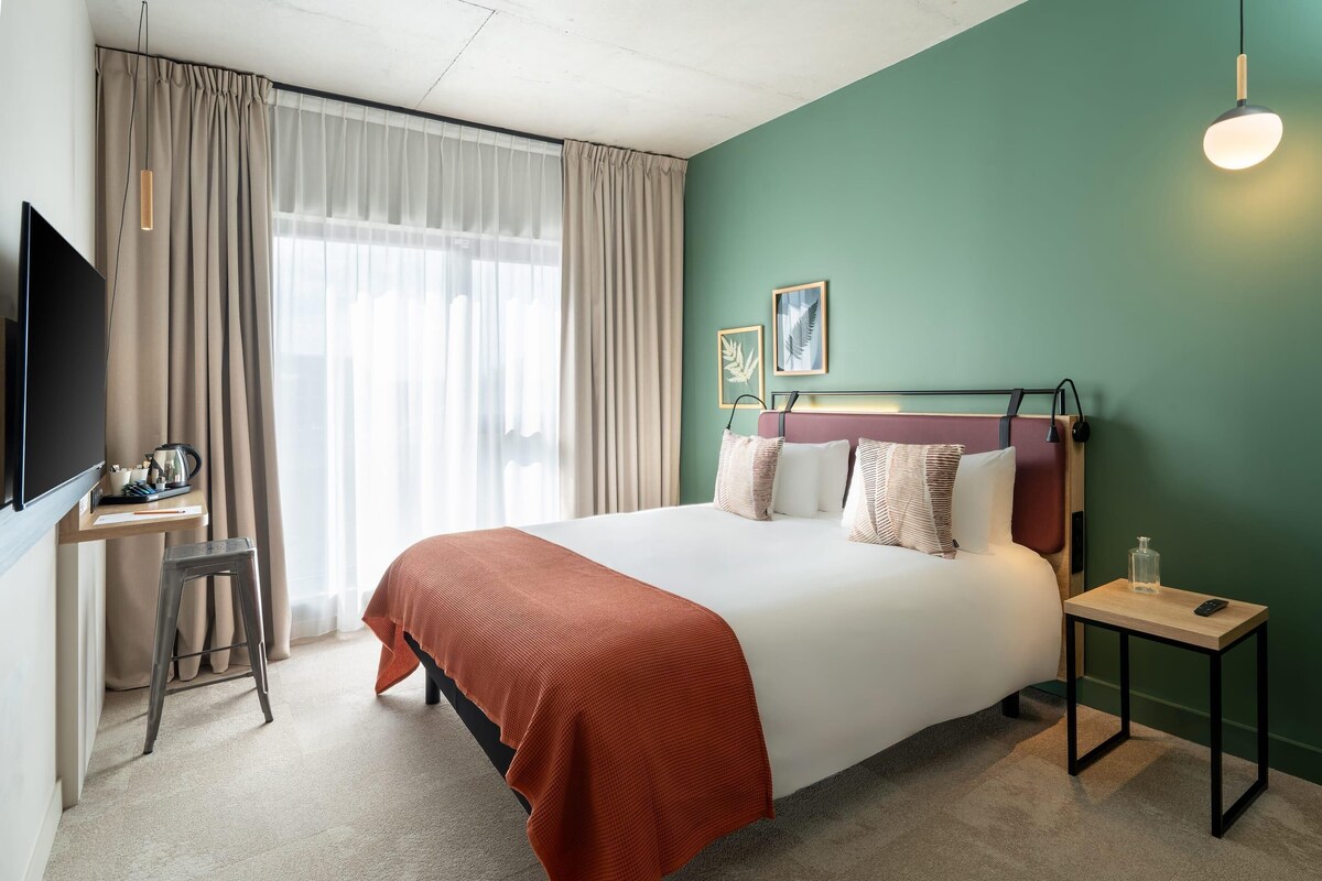 A hotel room with a bed, a desk, a chair, a TV, a bedside table, and a lamp.