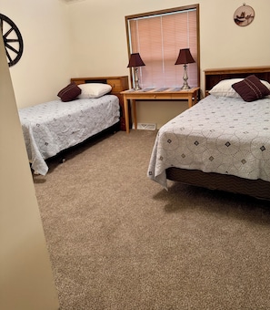 A room with two beds, a wooden nightstand, a window with blinds, and a wall-mounted lamp.