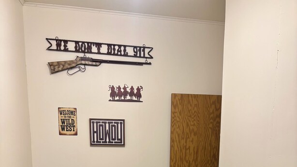 Ein Waschraum mit einer Sitzbank, einer Waschmaschine und Wanddekorationen, darunter Schilder mit den Aufschriften 'WELCOME TO THE WILD WEST' und 'HOW YOU'.