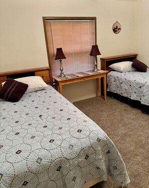 A bedroom with two beds, a nightstand, a table, and a wall-mounted lamp. - Spud Hill Hideaway - Old West charm with modern comfort (Lead)