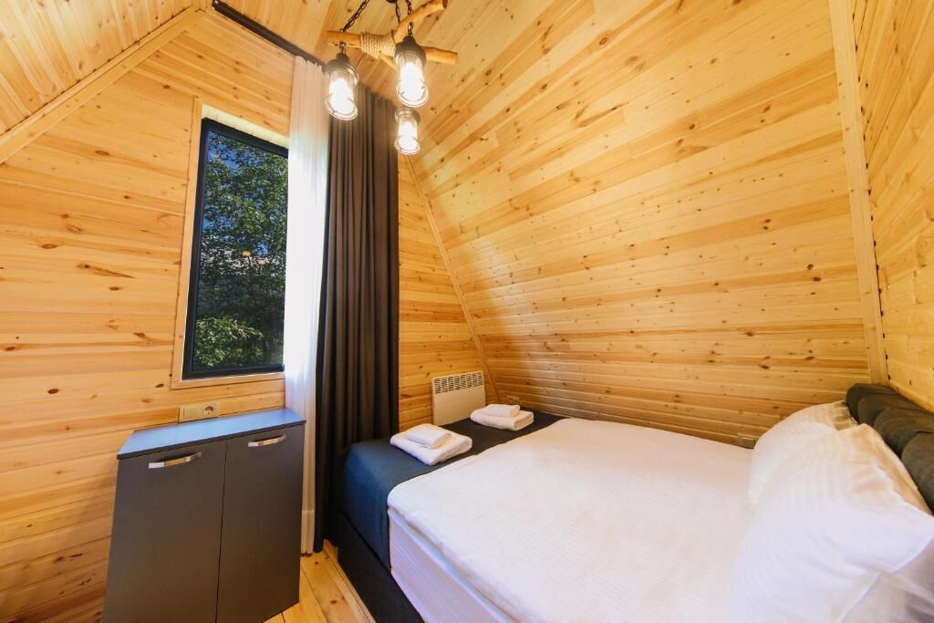 Quarto de cabana de madeira com cama, um pequeno armário, janela com cortinas e luminária no teto.