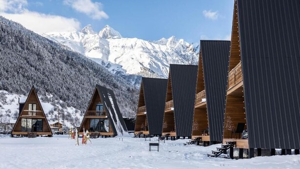 Een rij houten vakantiehuisjes met grote ramen, met een besneeuwde berg op de achtergrond.