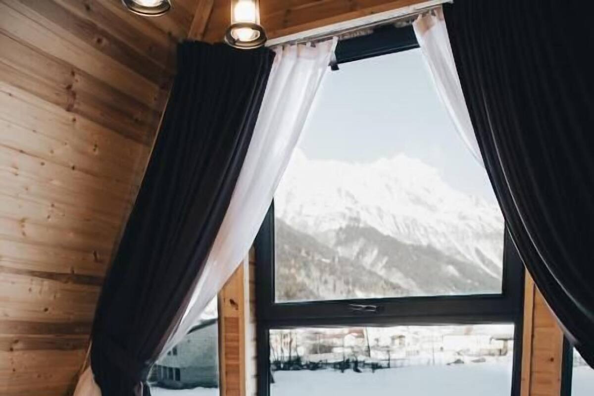 A wooden cabin room with a bed, a window with curtains, and a view of a mountain.