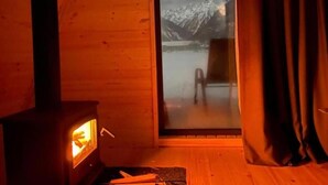 Een houten blokhut van binnen, met een houtkachel, een groot raam met uitzicht op besneeuwde bergen en een schuifdeur.