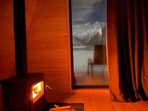 Interior de una cabaña de madera con estufa de leña, un ventanal con vistas a montañas nevadas y una puerta corrediza de vidrio.