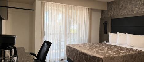 A hotel room with a bed, a desk with a coffee maker, a chair, a window with sheer curtains, and an air conditioning unit.