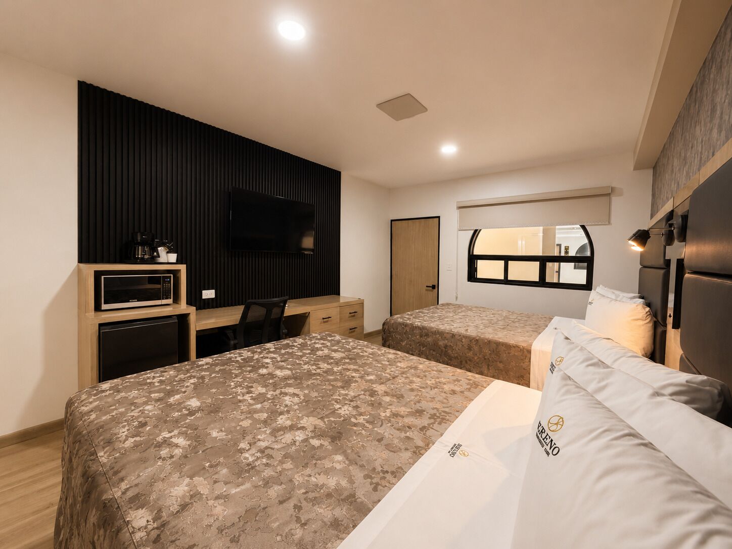 A hotel room with two beds, a desk, a TV, and a window.